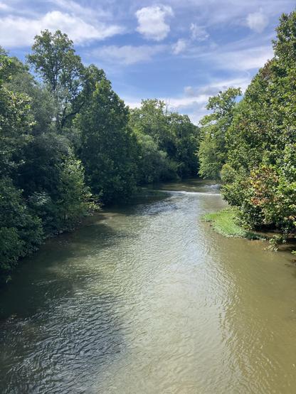 The now tranquil Roanoke River, flowing through lush greenery under a partly cloudy sky. Trees line both sides of the river, creating a serene and natural setting.