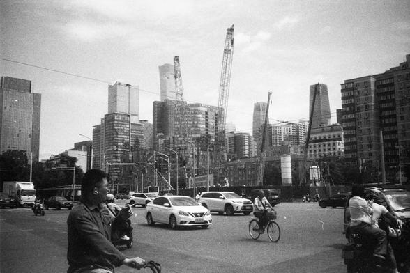 • English alt text: Black-and-white photo of a bustling city street with cars, motorcycles, and bicycles; people ride in the foreground while high-rise buildings and construction cranes dominate the background.
• 中文替代文字： 黑白照片展示繁忙的城市街道，前景有骑摩托车和自行车的人，背景是高楼和施工吊塔，展现城市快速发展的景象。