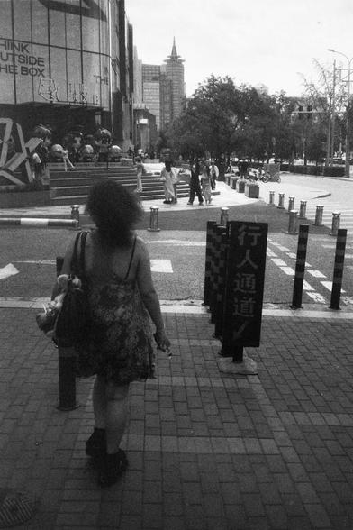 • English alt text: Black-and-white image of a person walking toward a pedestrian passage marked by bollards and a sign with Chinese characters; background includes people, a building with “THINK OUTSIDE THE BOX” signage, and urban elements.
• 中文替代文字： 黑白图像中，一人背对镜头走向标有“行人通道”的人行通道，背景有行人、写有“THINK OUTSIDE THE BOX”的建筑、雕塑和城市景观。