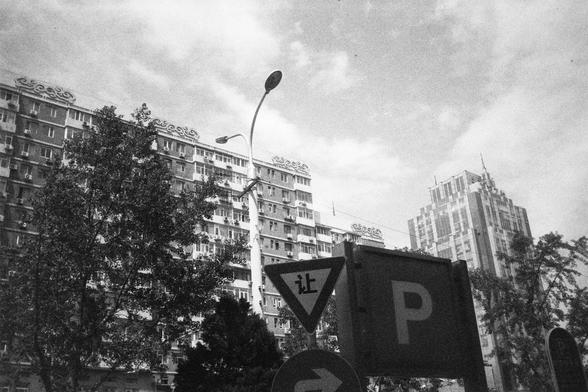 • English alt text: Black-and-white cityscape featuring tall buildings with ornate architecture, trees, a streetlight, and traffic signs including a Chinese yield sign and a parking sign.
• 中文替代文字： 黑白城市景观，背景是高楼，其中一栋顶部装饰精美；前景有树木、路灯，以及交通标志，包括一个写有中文的让行标志和一个“P”停车标志。