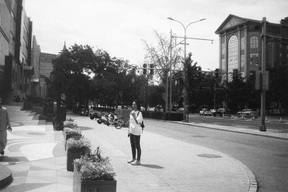 • English alt text: Black-and-white photo of a city street showing a person with a backpack standing on a tiled sidewalk near planters, with buildings, parked motorcycles, bicycles, cars, and pedestrians in the background.
• 中文替代文字： 黑白城市街景照片，一人背包站在人行道上，周围有花坛、建筑物、停放的摩托车和自行车，街道上有行人和行驶的汽车，展现城市日常生活场景。
