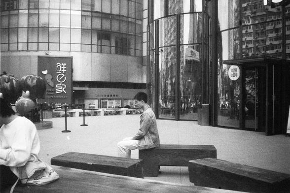 English Alt Text:
Two people sit on wooden benches in front of a modern glass building. One wears a white sweater and looks down, while the other wears a denim jacket and white pants. A sign reads “Fresh Food Supermarket,” and large apple sculptures decorate the plaza.

中文替代文字：
两个人坐在现代玻璃建筑前的木质长椅上，一人穿白色毛衣低头看东西，另一人穿牛仔夹克和白裤子。背景中有“Fresh Food Supermarket”的招牌和几个大型苹果雕塑，场景位于一个公共广场。