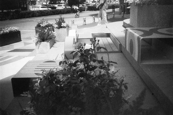 English Alt Text:
A black-and-white photo of an urban outdoor space with geometric benches, planters filled with flowers, and a checkerboard seating area. A person walks on the sidewalk in the background near parked cars.

中文替代文字：
一张黑白照片展示城市户外空间，有几何形状的长椅、种满花草的花坛，以及一个棋盘格图案的座位区。背景中有人在人行道上行走，旁边停着几辆车。