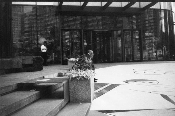 English Alt Text:
A person stands near the entrance of a modern building with large glass windows and a revolving door, while another sits on steps beside a flower planter. Abstract shapes and faces are drawn on the ground, and reflections of nearby buildings appear in the glass.

中文替代文字：
一个人站在带有旋转门和大玻璃窗的现代建筑入口处，另一个人坐在花坛旁的台阶上。地面上绘有抽象图形和面孔，玻璃窗中映出周围建筑的倒影。