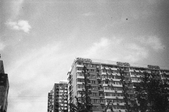 English Alt Text:
A black-and-white cityscape showing several multi-story residential buildings with balconies and air conditioning units. The building on the right features decorative trim along the roofline. The sky is mostly clear with a few scattered clouds.

中文替代文字：
一张黑白城市风景照，展示几栋多层住宅楼，带有阳台和空调设备。右侧的建筑屋顶边缘有装饰性设计。天空晴朗，只有少量云朵