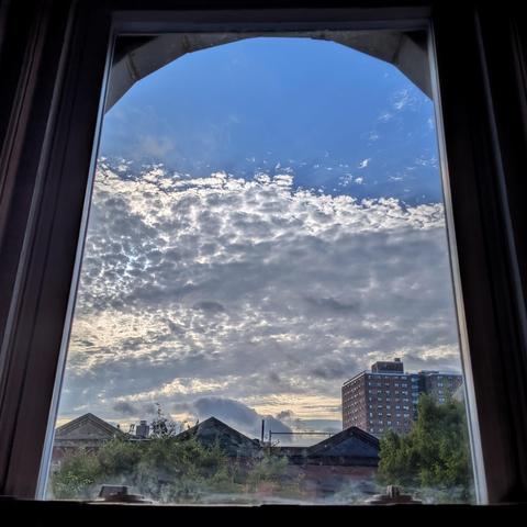 Looking through an arched window two hours after sunrise the bottom two-thirds of the blue sky is filled with a dense layer of white and grey clouds. Pointed roofs of Harlem brownstones are silhouetted across the street, and a taller apartment building can be seen in the distance. The green tops of two trees are on the bottom and right.