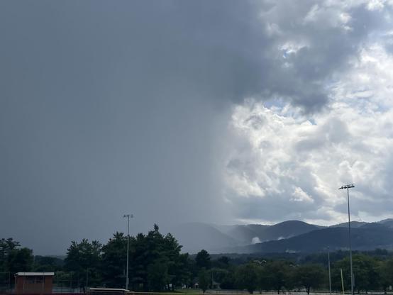 A dramatic sky displaying contrasting weather conditions: dark storm clouds and bands of rain on the left and lighter, partially cloudy skies on the right over distant mountains. Trees and sports field lights are visible in the foreground.