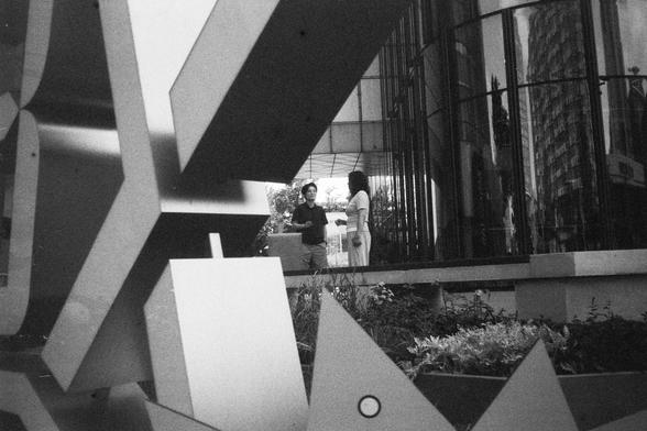 English Alt Text:
Black-and-white photo of two people talking near a modern building with reflective glass. A large abstract sculpture made of geometric shapes stands in the foreground. A patterned tall building is visible through the glass in the background.

中文替代文字：
黑白照片中，两人站在现代玻璃建筑前交谈。前景是一座由几何图形组成的大型抽象雕塑。背景玻璃后可见一栋带有独特图案的高楼。
