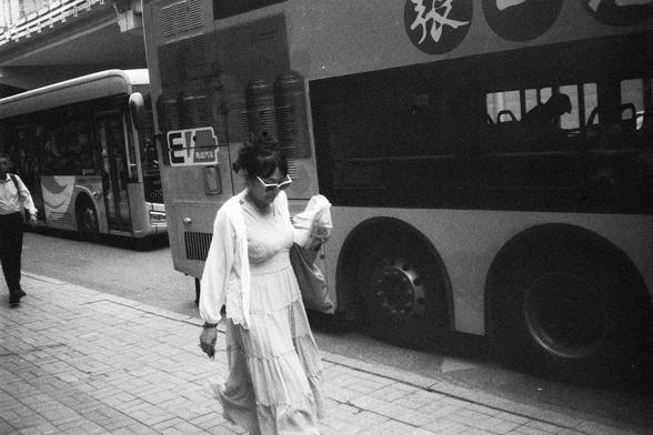 English Alt Text:
Black-and-white image of a city street with a double-decker and a single-decker bus. Two pedestrians walk along the sidewalk; one wears a long dress and carries a bag, the other wears a shirt and pants. The double-decker bus has Chinese characters and an “EV” sign.

中文替代文字：
黑白城市街景，一辆双层巴士和一辆单层巴士停靠在路边。两位行人走在人行道上，一位穿长裙提包，另一位穿衬衫和裤子。双层巴士上有“EV”标志和中文文字。