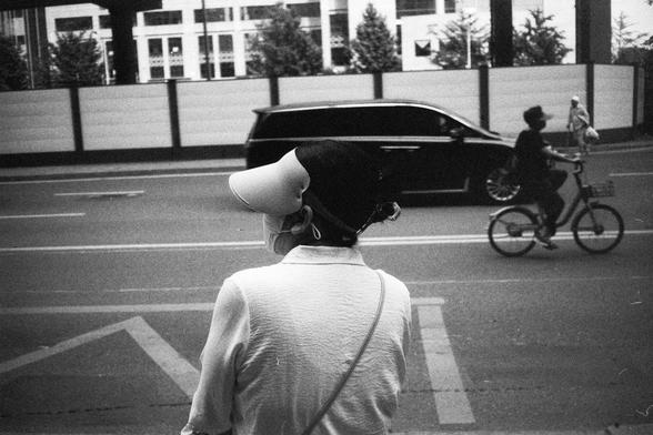 English Alt Text:
Black-and-white photo of a person wearing a visor and face mask, seen from behind while standing on a sidewalk. In the background, a car drives by, a cyclist rides along the road, and another pedestrian walks near a building with trees and a fence.

中文替代文字：
黑白照片中，一位戴遮阳帽和口罩的人从背后拍摄，站在人行道上望向街道。背景中有一辆汽车驶过，一人骑车经过，另一人沿着有树木和围栏的建筑旁行走。
