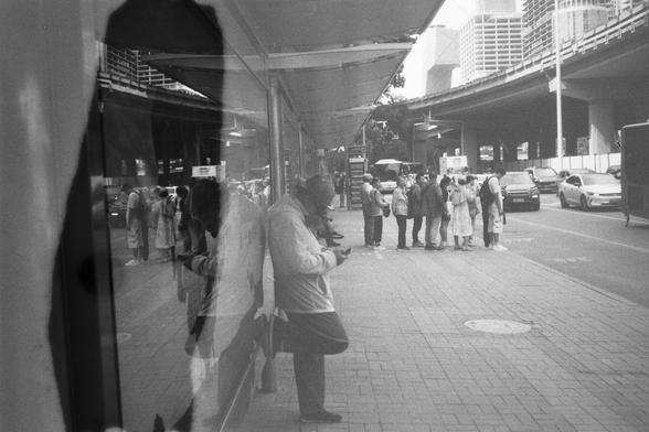• English Alt Text: Black and white photo of a busy urban street with people standing and conversing on the sidewalk, one person leaning against a wall using a phone, and reflections in glass adding depth; cars, buses, and buildings visible in the background.
• 中文替代文字： 黑白街景照片，几位行人站在人行道上交谈，一人靠墙看手机，玻璃反射增添层次感；背景中有汽车、公交车和建筑物。