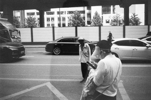 • English Alt Text: Black and white image of a street with a truck, a black car, and a white car; two people in the foreground, one wearing a hat and standing, the other wearing a cap and mask while looking at a phone; buildings and trees behind a fence in the background.
• 中文替代文字： 黑白街道照片，左侧有卡车，中间是黑色轿车，右侧是白色轿车；前景中两人，一人戴帽站立，另一人戴鸭舌帽和口罩看手机；背景有建筑和树木，被围栏隔开。