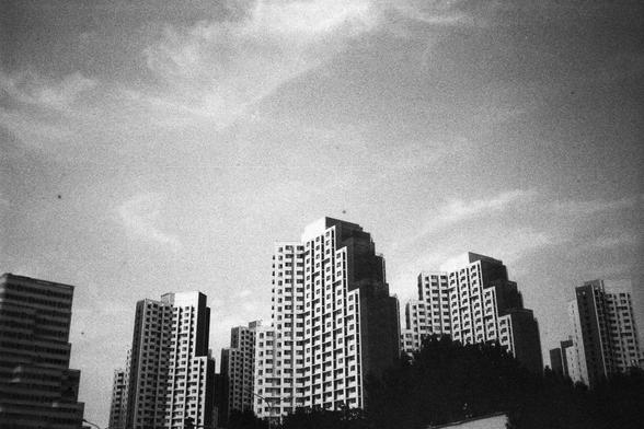 • English Alt Text: Black and white photograph of modern high-rise buildings with stepped architecture and many windows, set against a partly cloudy sky.
• 中文替代文字： 黑白照片展示现代高层建筑，结构呈阶梯状，窗户众多，背景是部分多云的天空。
