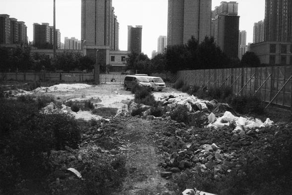 • English Alt Text: Black and white photo of an urban lot filled with rubble, debris, and overgrown plants; two vehicles are parked in the center, with tall buildings in the background, a fence on the right, and trees on the left.
• 中文替代文字： 黑白城市空地照片，地面布满瓦砾、杂物和杂草，中间停着两辆车；背景是高层建筑，右侧有围栏，左侧是树木。