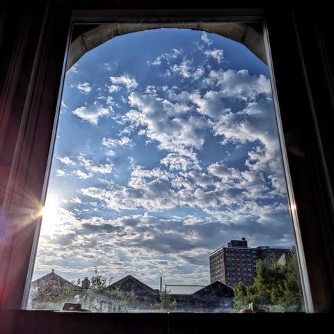Looking through an arched window ninety minutes after sunrise the blue sky is filled with fractal paisley white clouds. Pointed roofs of Harlem brownstones are silhouetted across the street, and a taller apartment building can be seen in the distance. The green tops of two trees are on the bottom and right.