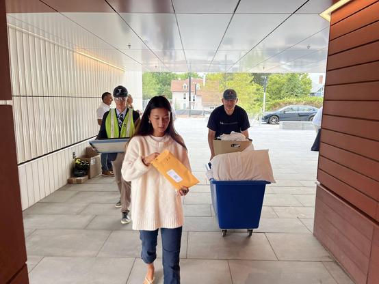 A student with an envelope followed by two men carrying items.