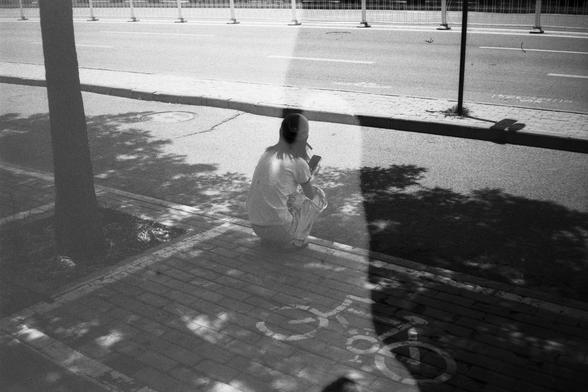• English Alt Text:
Black-and-white image of a person squatting on a brick sidewalk with a painted bicycle symbol, possibly eating or drinking. A ghostly double exposure effect and tree shadows create a moody, artistic scene.
• 中文替代文字：
黑白图像，一人蹲在铺有砖块并标有自行车符号的人行道上，似乎在吃或喝东西。图像中有树影和幽灵般的双重曝光效果，营造出一种情绪化的艺术氛围。