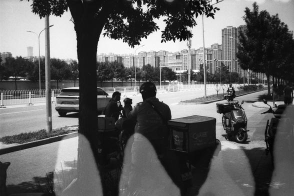 • English Alt Text:
Black-and-white photo of two delivery riders on motorcycles parked under a tree on a city sidewalk. One box shows the logo “闪送.” A bicycle rider and a car are visible on the street, with tall buildings in the background.
• 中文替代文字：
黑白照片，城市人行道上两名快递员骑摩托车停在树荫下，其中一个快递箱上印有“闪送”标志。街道上有骑自行车的人和一辆汽车，背景是高楼建筑。