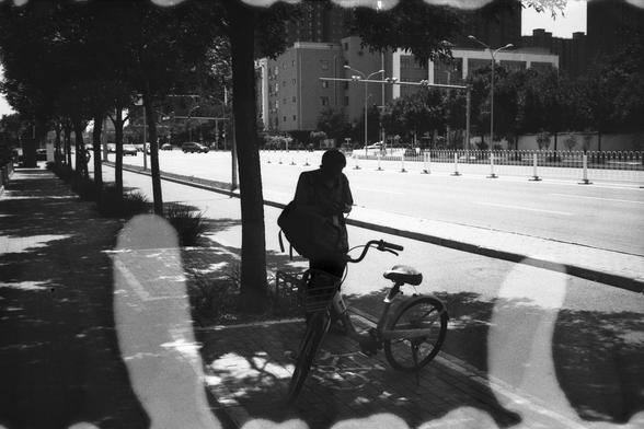 • English Alt Text:
A black-and-white photo of a person standing beside a bicycle on a tree-lined sidewalk, facing an empty road. Abstract white shapes from film damage or double exposure add a surreal, artistic effect.
• 中文替代文字：
一张黑白照片，画面中一人站在树荫人行道上的自行车旁，面朝空旷的马路。图像边缘和中央有抽象白色图形，可能是胶片损坏或双重曝光造成，增添了超现实的艺术感。