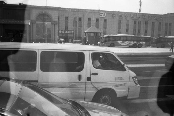 • English Alt Text:
A black-and-white photo of a city street in front of the “Da Jiao Ting International Business Hotel.” Several vehicles, including a white van labeled “Maximum capacity: 10 people,” a bus, and a car, are parked outside. Reflections and silhouettes suggest the photo was taken through a window.
• 中文替代文字：
黑白照片，城市街道场景，位于“大郊亭国际商务酒店”前。酒店外停有多辆车，包括一辆标有“准乘10人”的白色面包车、一辆公交车和一辆轿车。图像中有反光和人影，显示照片可能是透过窗户拍摄的。
