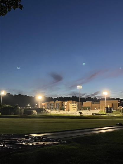 A softly illuminated sports field at dusk, featuring stadium lights and a school building in the background. The sky is darkening with wispy clouds, and a crescent moon is faintly visible. A tarp is laid on the ground in the foreground.