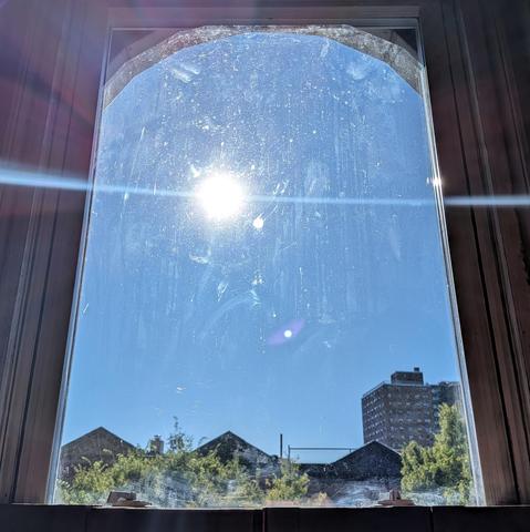 Looking through an arched window three hours after sunrise the sun is in the middle of the cloudless blue sky, creating a dramatic lens flare which bisects the frame horizontally in a curved white line. The direct sunlight is highlighting dirt and scratches on the glass of the window. Pointed roofs of Harlem brownstones are silhouetted across the street, and a taller apartment building can be seen in the distance. The green tops of two trees are on the bottom and right.