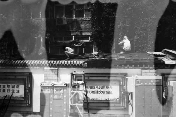 English Alt Text:
A black and white double exposure photograph showing two people working on a rooftop, overlaid with a wall featuring Chinese signage and utility boxes. The composition blends manual labor with urban infrastructure, creating a layered, artistic cityscape.

中文替代文字：
一张黑白双重曝光照片，画面中两人在屋顶上工作，叠加了一面带有中文标语和公共设施箱的墙面。画面融合了体力劳动与城市基础设施，呈现出层次丰富的艺术城市景象。