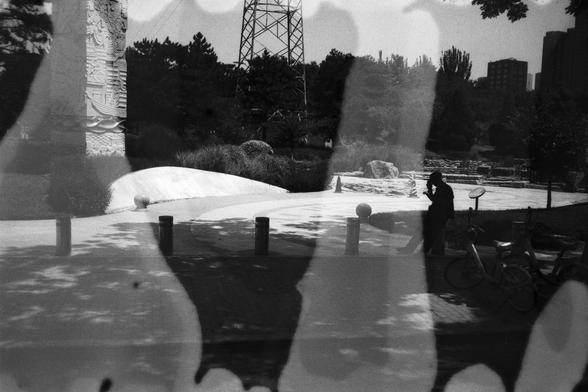 English Alt Text:
A surreal park scene viewed through a reflective surface, showing trees, rocks, a metal tower, and a person with a bicycle. Abstract dark shapes overlay the image, adding depth and visual complexity.

中文替代文字：
通过反光表面拍摄的超现实公园景象，画面中有树木、岩石、金属塔以及一位骑自行车的人。图像上覆盖着抽象的暗影形状，增强了画面的深度和视觉复杂性。