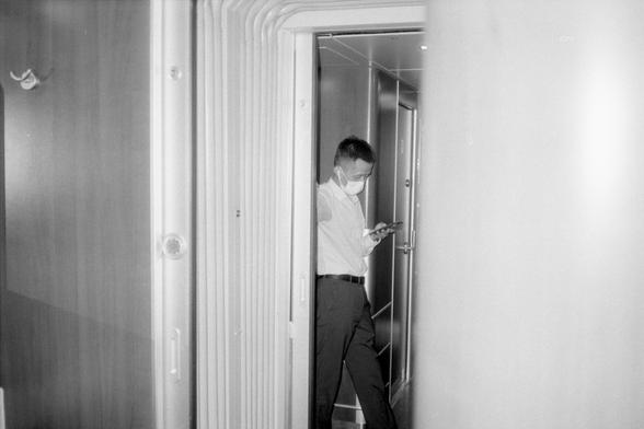 Kentmere Pan 200 (FF)

English Alt Text:
Black and white photo of a person standing in a narrow train corridor, wearing a face mask and looking at a smartphone. The person is dressed in a light shirt and dark pants. The corridor has metallic walls and doors, creating a confined indoor atmosphere.

中文替代文字：
黑白照片，一人站在狭窄的列车走廊中，戴着口罩，低头看手机。此人穿着浅色衬衫和深色裤子。走廊两侧是金属墙和门，营造出封闭的室内环境。
