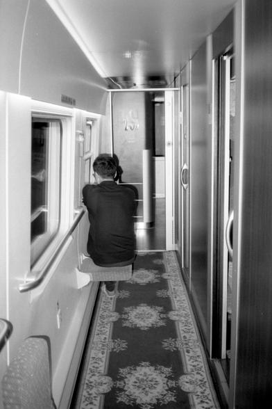 Kentmere Pan 200 (FF)

English Alt Text:
Black and white image of a person seated on a fold-down wall seat in a narrow train corridor. The person faces away from the camera, looking toward the end of the hallway. The corridor has patterned carpet, windows on the left, and doors on the right.

中文替代文字：
黑白图像，一人坐在列车走廊中靠墙的折叠座椅上，背对镜头，望向走廊尽头。地板铺有图案地毯，左侧是窗户，右侧是车厢门。