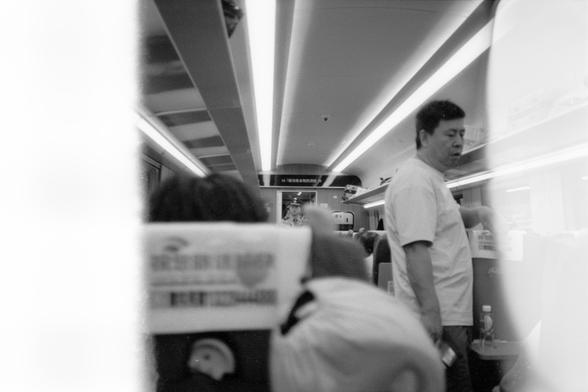 Kentmere Pan 200 (FF)

English Alt Text:
Black and white photo taken from a seated passenger’s perspective inside a train. The view looks down the aisle, showing overhead lights and luggage compartments. Several passengers are visible, including a man standing in the aisle wearing a light shirt. Seat headrests have white covers with Chinese text. The image has light leaks on both edges, creating a faded effect.

中文替代文字：
黑白照片，从乘客座位视角拍摄，展示列车过道景象。画面中有顶部灯光和行李架，几位乘客可见，其中一名男子穿浅色衬衫站在过道中。座椅头枕上覆盖着印有中文的白…