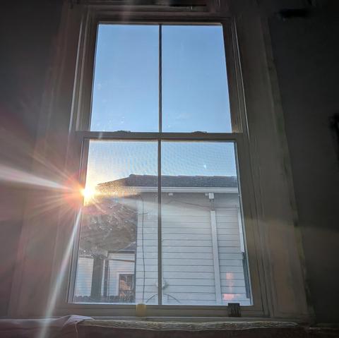 Looking through a window ninety minutes after sunrise the blue sky is cloudless. The sun is rising over a clapboard house, creating a dramatic lens flare. 
