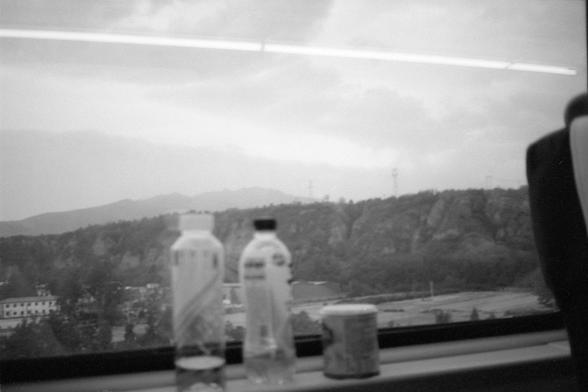 Kentmere Pan 200 (FF)

English Alt Text:
A black-and-white photo taken from inside a moving train, showing a mountainous landscape with rocky cliffs, power lines, and sparse vegetation. Three beverage containers sit on the window ledge in the foreground, slightly blurred due to motion.

中文替代文字：
一张黑白照片，从行驶中的火车车窗内拍摄，窗外是山地景观，岩石峭壁、电线和稀疏植被。前景中窗台上放着三个饮料容器，因运动而略显模糊。