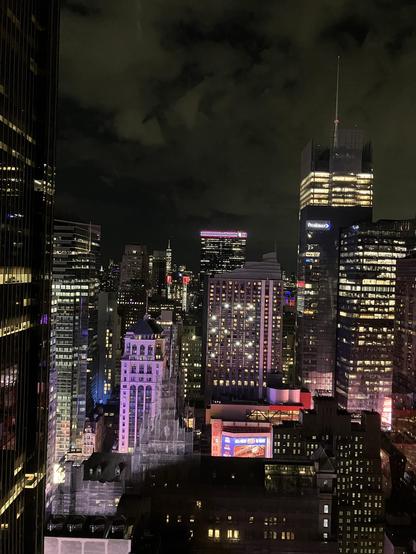 A nighttime view of the Manhattan skyline, showcasing tall buildings illuminated with colorful lights. Features include a historic building with a distinctive rooftop, modern skyscrapers, and a backdrop of clouds partially obscuring the sky.