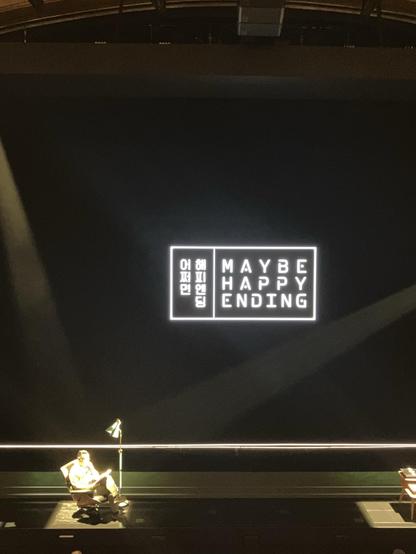 A stage with a dark background featuring the text "MAYBE HAPPY ENDING" in illuminated letters. A person is seated in a chair with a lamp, reading. A small table is nearby.