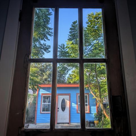 Looking through a six-panes window two hours after sunrise the sky is blue and cloudless. A small wood house is painted almost the same color as the sky, with orange trim. The front door of the small house is white with an oval window. Tall gangly trees surround the house.