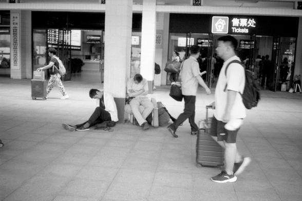 Kentmere Pan 200 (FF)

• English Alt Text:
A black-and-white image of a train station interior. Three people sit or lie on the ground near a pillar with luggage, while others walk by with suitcases. A ticket counter with bilingual signage is visible in the background.
• 中文替代文字：
一张黑白照片，拍摄于火车站室内。三人靠近柱子坐着或躺着，旁边有行李，其他人拖着行李箱走过。背景中可见一个售票窗口，标有中英文标识。