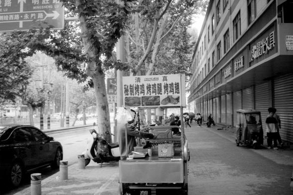 Kentmere Pan 200 (FF)

English Alt Text:
A black-and-white photo of a Chinese sidewalk with a metal food cart in the foreground. The cart has a canopy and displays Chinese signs with images of street food. Scooters and a car are parked along the curb behind it. Trees line the sidewalk, casting shade. Further down, pedestrians walk past shuttered storefronts with Chinese signage. The scene feels local and lived-in, capturing the rhythm of everyday life.

中文替代文本：
黑白照片中，一辆金属小吃车停在人行道上，车顶有遮阳棚，挂着中文招牌…