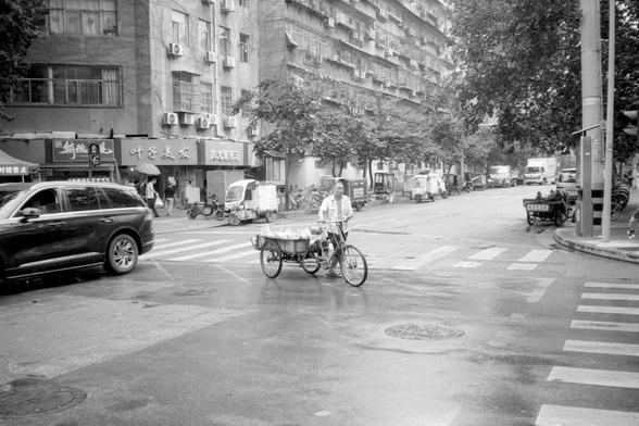 Kentmere Pan 200 (FF)

English Alt Text:
A black-and-white street scene shows a person pushing a tricycle cart across a crosswalk. The cart has a large front container, likely used for deliveries or goods. A black SUV waits nearby. The street is lined with scooters, cars, and a multi-story building covered in air conditioners and Chinese shop signs. Trees and pedestrians fill the background, creating a busy, everyday urban atmosphere.

中文替代文本：
黑白街景中，一人推着三轮车穿过斑马线，车前载物箱可能用于送货。旁边一辆黑色 SUV 停在路口。街道两侧…