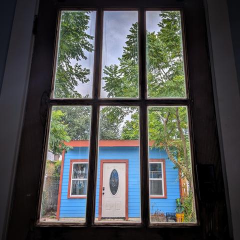 Looking through a six-paned window an hour after sunrise the sky is full of gray clouds. A small wood house is painted bright blue with orange trim. The front door of the small house is white with an oval window. Tall gangly trees surround the house.