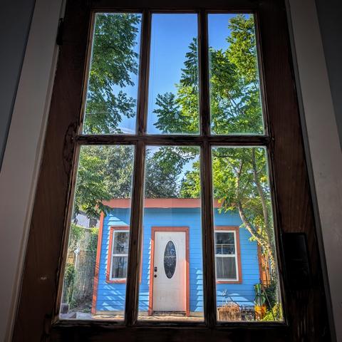 Looking through a six-paned window two hours after sunrise the sky is bright blue with a few stringy white clouds. A small wood house is painted almost the same color as the sky, with orange trim. The front door of the small house is white with an oval window. Tall gangly trees surround the house.