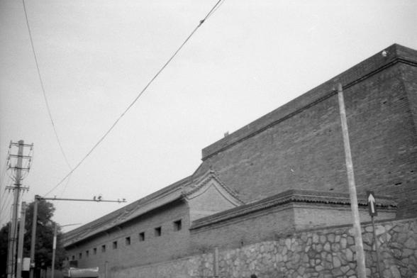 Kentmere Pan 200 (FF)

English Alt Text:
A long, traditional-style brick building stretches across the frame. Its base is made of stone, and the roof is tiled with a gentle slope and ornate eaves, suggesting historical or cultural significance. Small, evenly spaced windows line the upper wall, and utility poles with wires run parallel to the street beside it. A traffic sign stands on the right. The black-and-white tone gives the image a vintage feel, evoking a quiet, bygone era.

中文替代文字：
画面中是一座…