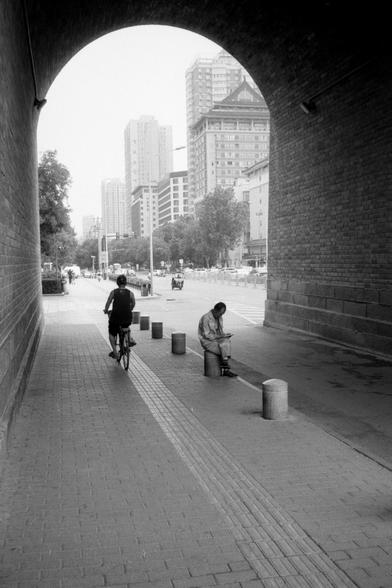 Kentmere Pan 200 (FF)

English Alt Text:
A city scene viewed from inside a brick tunnel with a curved ceiling. Outside, the street is lined with tall buildings, trees, and vehicles. Inside the tunnel, a person rides a bicycle away from the camera, while another sits on a concrete bollard, reading or looking at something in their hands. The ground is paved with bricks and includes a tactile strip for visually impaired pedestrians. The contrast between the shadowed tunnel and the bright city beyo…