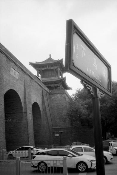 Kentmere Pan 200 (FF)

English Alt Text:
A black-and-white photo of a traditional Chinese gatehouse with a pagoda-style roof, built atop a brick city wall. The structure includes arched entrances and is surrounded by parked cars and trees. In the foreground, a modern street sign reads “XIAMALING” in Chinese and English. On the wall, the characters “文昌門” are visible, suggesting a historical or cultural site. The image blends old and new, showing urban development alongside preserved architecture…