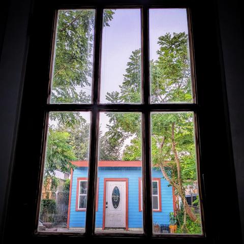 Looking through a six-paned window eight minutes before sunrise the sky is dusty blue. A small wood house is painted bright blue with orange trim. The front door of the small house is white with an oval window. Tall gangly trees surround the house.