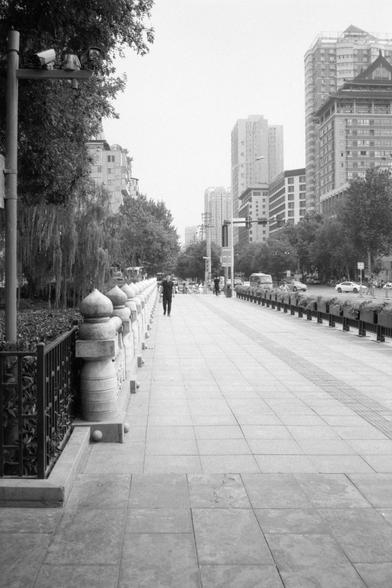 Kentmere Pan 200 (FF)

English:
A black-and-white photo of a city sidewalk. Stone posts line the left, and a metal railing borders the right. Tall modern buildings rise on both sides, with mixed architectural styles. A few pedestrians walk along the path, and cars drive down the road. Trees add greenery, blending nature with urban life.

中文：
黑白城市人行道照片。左侧是石柱，右侧是金属栏杆。两旁是高大的现代建筑，风格多样。几位行人沿路行走，车辆在道路上行驶。树木点缀其间，自然与城市融合。