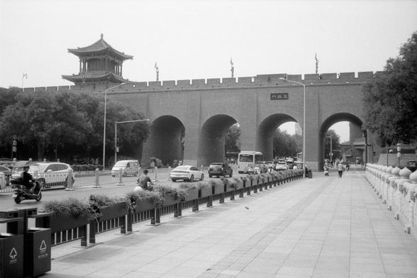 Kentmere Pan 200 (FF)

English:
A grayscale image of a traditional Chinese city wall with arched gateways. A watchtower with tiered roofs sits atop the wall. The central arch bears the sign “文昌門”. Cars, buses, and scooters pass through the arches, merging ancient architecture with modern traffic. Trees and planters line the walkway.

中文：
灰度图像，展示一座传统中国城墙，设有多个拱门。城墙上方是带有飞檐的瞭望塔。中央拱门上写着“文昌門”。汽车、公交车和摩托车穿行其间，古代建筑与现代交通交织。左侧有树木，右侧人行道设有花坛和护栏
