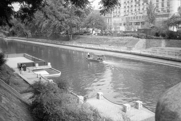 Kentmere Pan 200 (FF)

English:
A peaceful canal scene in monochrome. A small boat moves through the water, likely for maintenance. A person stands near the canal’s edge. Decorative stone posts and railings line the walkway. Trees and buildings, including one with a grid-like facade, form the backdrop.

中文：
宁静的城市运河场景，黑白色调。一艘小船在水中行驶，可能用于维护。一个人站在运河边。人行道上有装饰性石柱和栏杆。背景是树木和多层建筑，其中一栋外墙呈网格状。