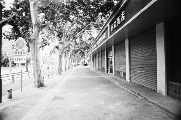 Lucky Lucky SHD 400 (FF)

🔹 English Alt Text

A quiet sidewalk scene with trees lining the left side, casting shade. On the right, closed storefronts with metal shutters and signage in Chinese, including “百家商店” (Hundred Family Store), suggest a general goods shop. Tactile paving runs along the brick sidewalk for visually impaired pedestrians. A circular traffic sign and a rectangular street sign are visible. The atmosphere is calm and subdued, enhanced by the black-and-white tones.

🔹 中文替代文字

一…