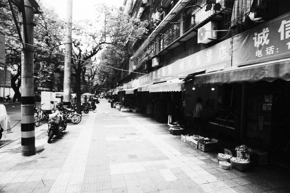 Lucky Lucky SHD 400 (FF)

🔹 English Alt Text

A narrow tiled street lined with market stalls and small shops. On the right, baskets of fruit—likely apples—are placed on the ground in front of awnings and signs with Chinese characters. On the left, motorcycles and bicycles are parked. Tall buildings with balconies and air conditioners loom above, and trees line both sides. The scene suggests a mixed-use neighborhood, blending residential and commercial life. The monochrome palette adds a nostalg…